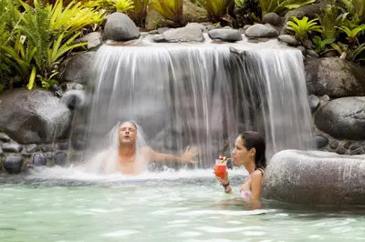 a man and a woman sitting in a swimming pool next to a waterfall at Hotel Arenal Springs Resort & Spa in Fortuna