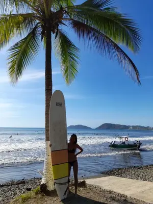 a man standing next to a surfboard next to a palm tree at BeachBungalow Surf&Stay in Patos