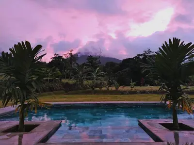 a pool with two palm trees in front of a sunset at Villa Luxury Volcano Paradise Retreat in Fortuna