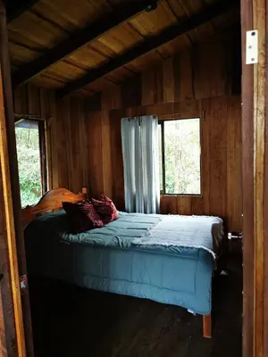 a bed in a wooden room with two windows at Rio Paquita Lodge in Tapantí