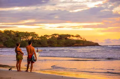 a man and woman standing on a beach with a surfboard at The Coast Beachfront Hotel in Tamarindo