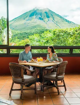 a man and woman sitting at a table with a mountain in the background at Hotel Arenal Kioro Suites & Spa in Fortuna