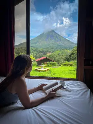 a woman laying on a bed reading a book and looking out a window at Arenal Roca Lodge & Bungalows in Fortuna