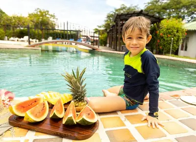 a young boy sitting next to a pool with a tray of fruit at Hotel El Rancho in Quepos