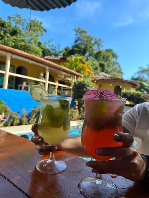 two hands holding two cocktails on a table near a pool at Hospedaje Colibri in Manuel Antonio