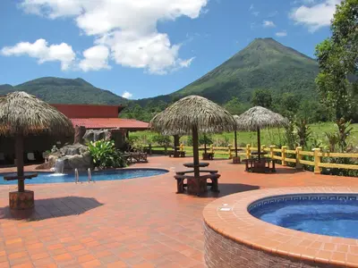a resort with a pool with tables and straw umbrellas at Hotel La Pradera del Arenal in Fortuna