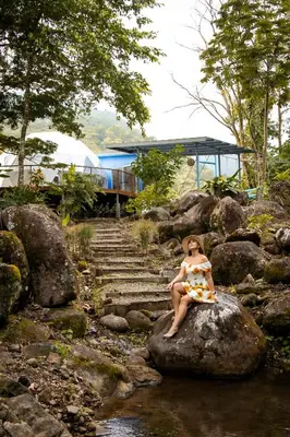 a woman in a dress sitting on a rock by a stream at Bitzu Dome-Bijagua Riverside Glamping in Bijagua